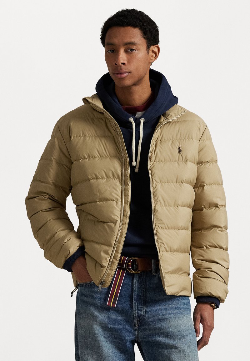 Beige pufferjakke med lynlås foran, hætte og quiltet design. Båret over en navy hoodie, parret med blå jeans og et brunt bælte.
