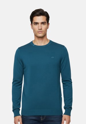Uomo che indossa un maglione a girocollo verde acqua con maniche lunghe e jeans scuri, in piedi contro uno sfondo chiaro e uniforme.