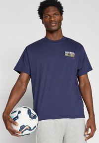 Camiseta de manga curta azul-marinho com o logótipo "Juventus Mille897" em branco e amarelo do lado esquerdo, acompanhada de uma bola de futebol com padrão.