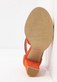 Sandale orange en daim à bout ouvert avec talon bloc, vue du côté de la semelle sur fond blanc, appuyée contre un mur.