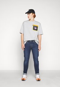 T-shirt cinza de manga curta com um bolso em malha acentuado em amarelo. Usada com jeans azul-escuro e ténis brancos com detalhes laranja. Boné preto.