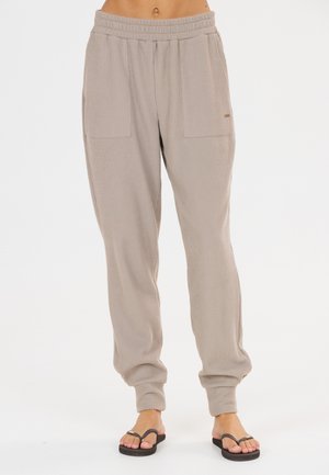 Beige geribde joggerbroek met elastische tailleband en boorden gedragen door een persoon met een blootstaande buik die blootsvoets staat met zwarte teenslippers.