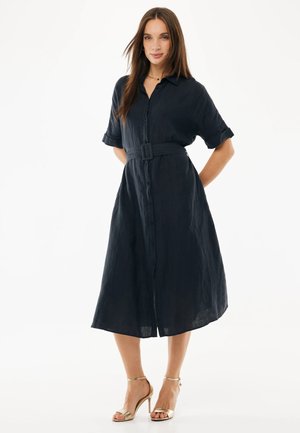 Žena ve středně modrých midi košilových šatech s páskem, krátkými rukávy a zlatými sandály na vysokém podpatku, stojící před bílým pozadím.