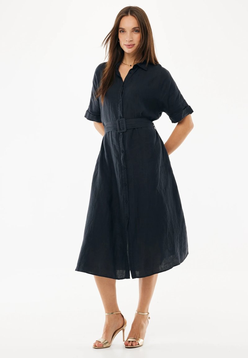 Kobieta ubrana w granatową midi sukienkę koszulową z paskiem, krótkimi rękawami i złotymi sandałami na wysokim obcasie, stojąca na białym tle.