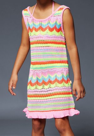 Dziecko w bezrękawowej, wielokolorowej dzianinowej sukience z wzorami zygzaków i pasków oraz różową falbaną na dole, stojące na szarym tle.