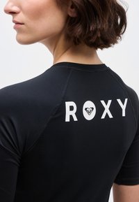 Γυναίκα με κοντά καστανά μαλλιά που φοράει μαύρο μπλουζάκι με λευκό λογότυπο "ROXY" και καρδιά στο πάνω μέρος της πλάτης, κοιτάζοντας πλάγια.