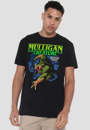Young man wearing black T-shirt with bright green and blue graphic of a monstrous creature and the text "Mulligan Creature" and "Stranger Things Tales 85."