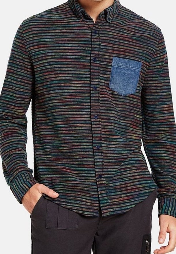 Homme portant une chemise à rayures multicolores avec des boutons, une poche poitrine en denim bleu et un pantalon noir, les mains dans les poches.