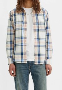 Chemise à carreaux avec des lignes bleues, vertes et orange sur fond blanc ; boutonnée à l'avant, manches longues, une poche poitrine. Portée sur un t-shirt blanc.