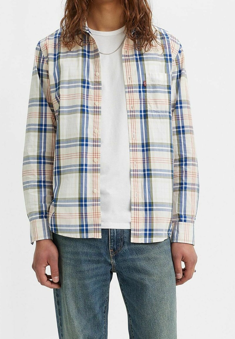 Chemise à carreaux avec des lignes bleues, vertes et orange sur fond blanc ; boutonnée à l'avant, manches longues, une poche poitrine. Portée sur un t-shirt blanc.