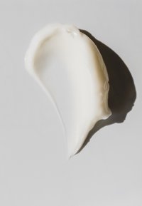 White cream texture with a glossy sheen, shaped like a curved drop, placed on a smooth white surface, highlighting its richness and consistency.