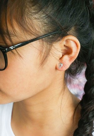 Silberner, delfinförmiger Ohrstecker, der in einem durchstochenen Ohr getragen wird, mit glatter, spiegelnder Oberfläche und filigranem Design. Schwarze randlose Brille sichtbar.