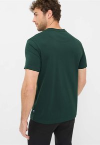 T-shirt verde scuro a maniche corte in cotone, con collo a girocollo e un piccolo dettaglio del logo all'orlo. Texture liscia.
