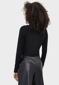 Kobieta z kręconymi włosami, ubrana w dopasowaną czarną bluzkę z długim rękawem i luźne czarne skórzane spodnie, pokazana od tylu na jednolitym tle.