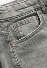 Close-up van grijs denimstof, met de focus op een zak, stikdetails in contrasterend bruin en een koperkleurige knoopaccent.