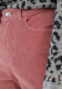 Roze corduroy broek met verticale ribbing, voorzien van een voorzak en een zilveren knoop in de tailleband, gecombineerd met een gebreid trui met patroon.