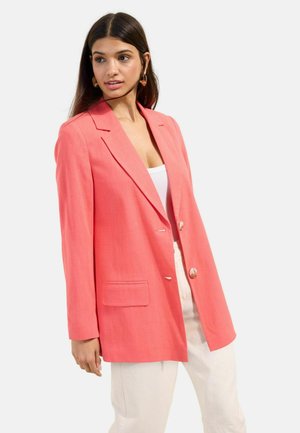 Woman wearing coral blazer over white top and light-colored pants, standing against a plain light background, looking to the side.