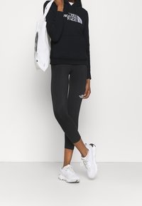 Schwarzer Hoodie mit weißem Logo, kombiniert mit schwarzen Leggings, die ein kleines Logo tragen, und weißen Turnschuhen. Die Tasche ist eine schlichte weiße Tragetasche.