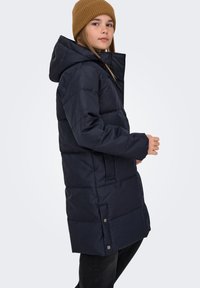 Temno modra puhovka s kapuco, s prešitim vzorcem, sprednjimi žepi in zapenjanjem na pritrdilne gumbe. Nošena čez črne hlače.