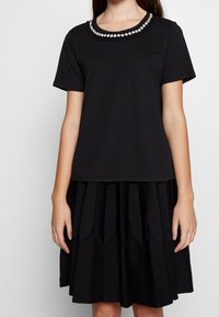 Svart bomullstopp med kort ärm och blomsterdekorerad halsringning, ihopparad med en svart plisserad kjol med ett texturerat mönster.