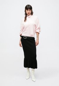 Lyserosa, strikket genser med sløyfedetalj, kombinert med en svart ribbestrikket skjørt med utslått kant. Hvite ankelstøvler fullfører antrekket.