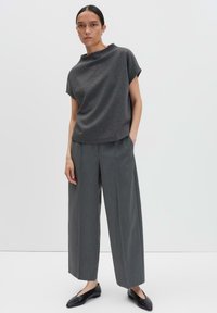 Vrouw in een donkergrijze top met korte mouwen, brede grijze broek en zwarte puntige ballerina's, staand tegen een effen witte achtergrond.