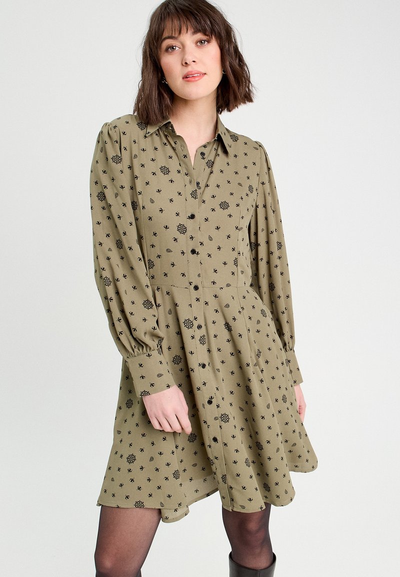 Robe vert olive avec un devant à boutons, des manches bouffantes longues et des motifs floraux noirs. Le tissu est léger et légèrement texturé.