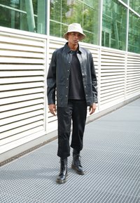 Chaqueta de camisa de cuero negra, camiseta negra, pantalones negros y botas con cordones negras con suela amarilla. El modelo lleva un sombrero de cubo color beige claro.