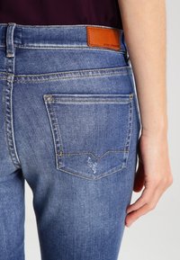 Ljust blå denimjeans med smal passform, som har en brun läderlogotyp, bakfickor och subtila slitningar.