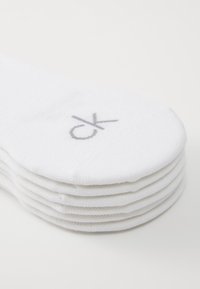 Stabel med fem hvite bomullssokker til ankler med grå "ck"-logo brodert nær tærne på en hvit bakgrunn.