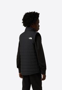 Čierna bezrukávová prešívaná vesta s prešívaným vzorom a vysokým golierom. Na ľavom ramene biele logo. Nosená cez čierne tričko s dlhým rukávom.