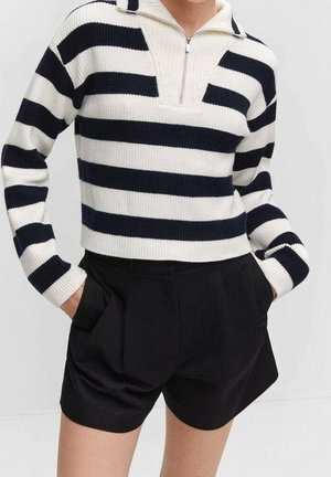 Personne portant un pull rayé large blanc et noir à manches longues avec un col à demi-zip, associé à un short noir taille haute avec des poches.