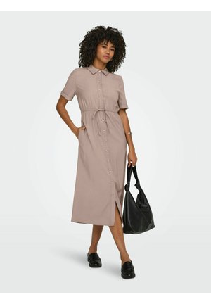 Kiharatukkainen nainen, jolla on päällään beige, nappikiinnitteinen midimekko, mustat loafers-tyyliset kengät ja musta käsilaukku, seisoo yksiväristä taustaa vasten.