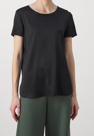 Personne portant un chemisier noir uni à manches courtes et un pantalon ample vert foncé sur un fond uni.
