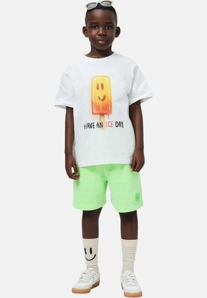 Witte katoenen T-shirt met een grafiek van een lachende ijslolly, gecombineerd met neon groene shorts, patroon sokken en witte sneakers met beige accenten.