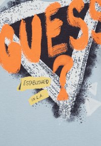 Camiseta de color azul claro con texto gráfico en negrita en naranja y negro, un diseño triangular y acentos amarillos que dice "Establecido en L.A."