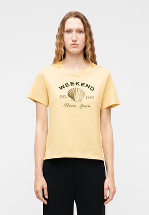 Ung person med langt bølget hår iført en gul T-shirt med grafik af en muslingeskal og teksten "WEEKEND EST. 1983 Ibiza, Spanien" samt sorte bukser.