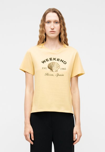 Jeune personne aux cheveux longs et ondulés portant un T-shirt jaune avec un motif coquillage et le texte « WEEKEND EST. 1983 Ibiza, Espagne » ainsi qu'un pantalon noir.