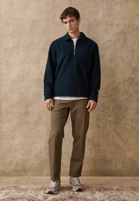 Tmavomodré polokošele s krátkym zipsom a prednou kapsou, v kombinácii s svetlohnedými nohavicami a šedými teniskami s bielymi akcentmi na vzorovanom koberci.