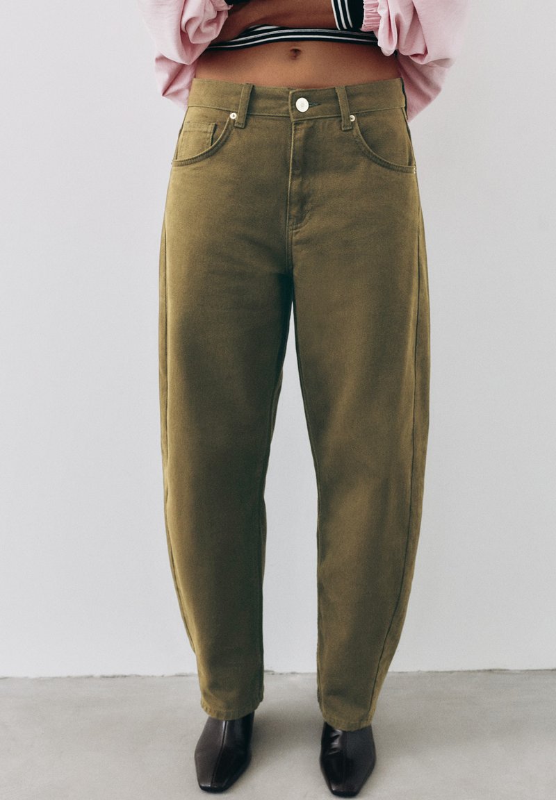 Personne portant un pantalon taille haute vert olive, des chaussures noires à bout carré, et un haut rose avec une ceinture rayée noire et blanche visible.
