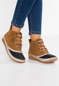 Bottines en daim marron avec embouts en caoutchouc noir, trois œillets pour les lacets et semelles en caoutchouc texturé, posées sur une surface blanche.