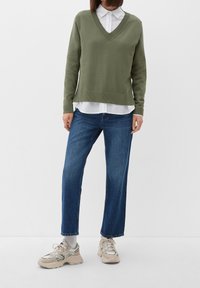 Olijfgroene V-hals trui, gelaagd over een witte blouse. Blauwe rechte jeans. Beige sneakers met meerkleurige accenten.