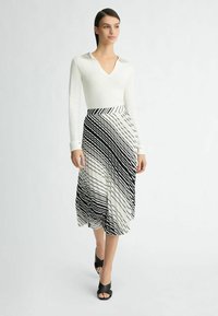 Blusa blanca de manga larga con cuello adornado, combinada con una falda plisada a rayas en blanco y negro, y sandalias de tacón abierto negras.