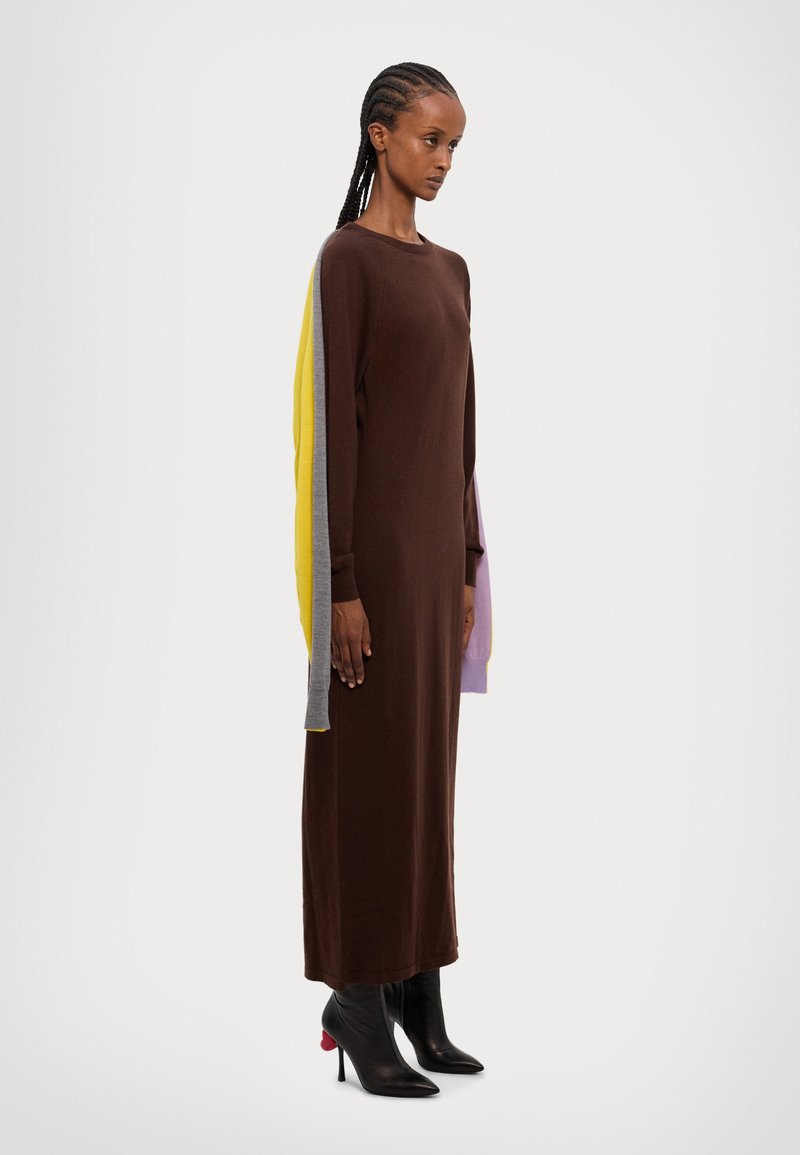Robe longue ajustée marron à manches longues, dans des teintes de jaune, gris et violet ; texture douce. Associée à des bottes noires à talons aiguilles.