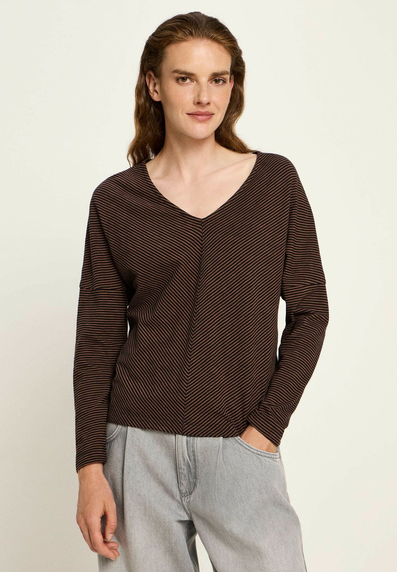 Woman wearing a brown and black thin-striped long-sleeve top with V-neck and light gray loose-fitting jeans, standing with one hand in pocket.