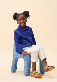 Niño que lleva una chaqueta de mezclilla azul, pantalones blancos y sandalias naranjas, sentado en una silla de plástico azul texturizada. Sonriendo, con el cabello rizado peinado en moños.