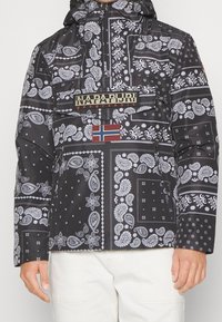 Zwarte jas met een patroon van witte paisleyprints, een klepzak met de tekst "Napapijri geographic" en een Noorse vlagpatch op de borst.