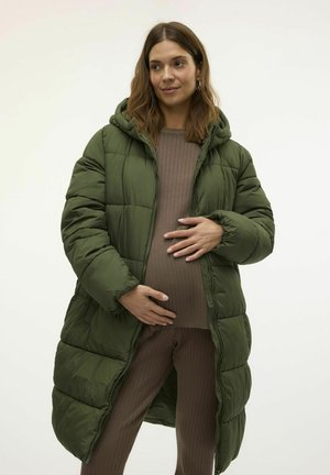 Manteau matelassé vert avec capuche, design capitonné et manches longues. Porté sur un haut brun côtelé et un pantalon assorti, mettant en valeur une silhouette de femme enceinte.