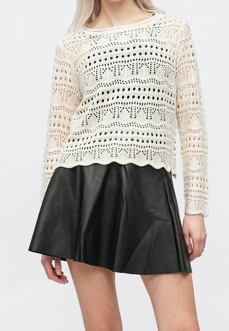 Femme portant un haut à manches longues en crochet crème et une jupe noire plissée en cuir, debout devant un fond blanc uni.