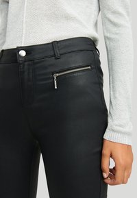 Svarta fusk läderbyxor med en silverfärgad dragkedjeficka, slät textur och en snäv design. Åtföljd av en ljusgrå tröja.